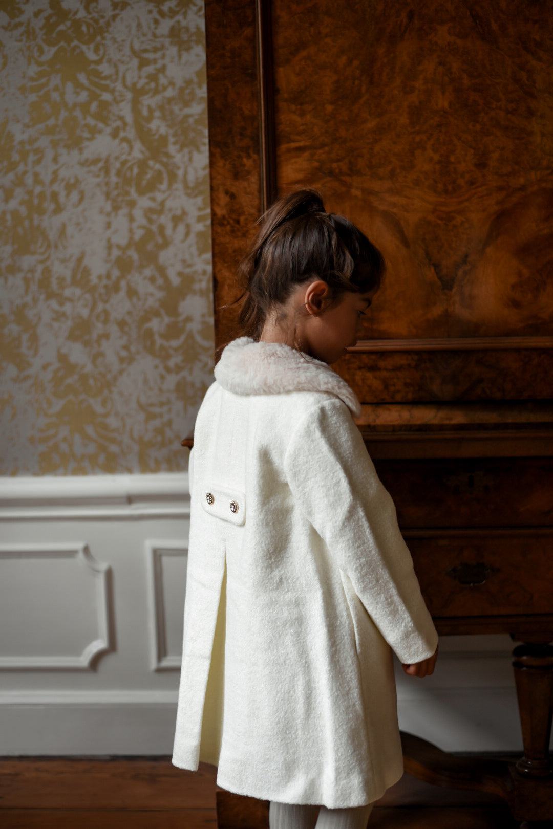 Back view of a girl wearing the Mara Tweed Coat in Ivory beside an antique wooden cabinet in the Canal House