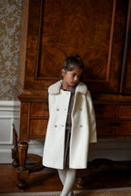 Girl wearing the Mara Tweed Coat in Ivory beside an antique wooden cabinet in the Canal House