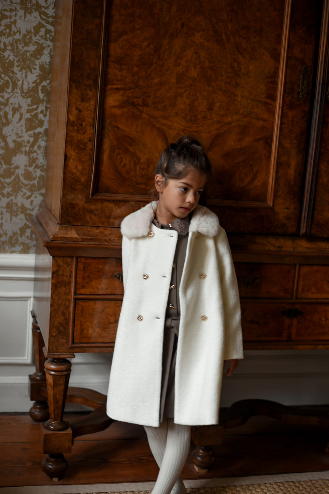 Girl wearing the Mara Tweed Coat in Ivory beside an antique wooden cabinet in the Canal House