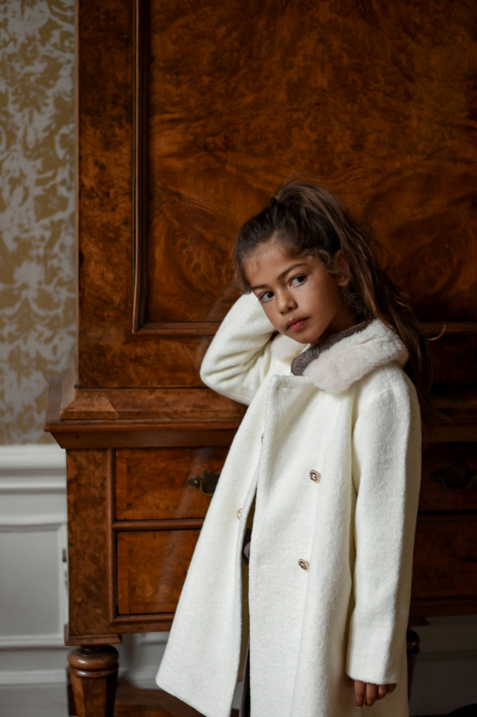 Front view of a girl wearing the Mara Tweed Coat in Ivory beside an antique wooden cabinet in the Canal House